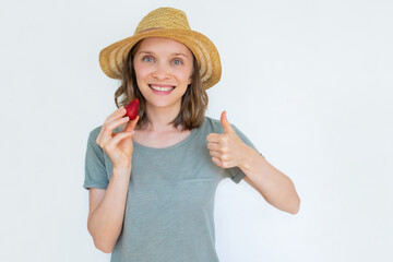 Smiling lady in hat holding ripe strawberry with thumb up and looking at camera. Portrait of young girl isolated on white background. Front view. Gesturing and nutrition concept