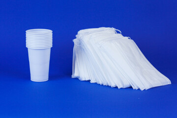 A stack of disposable medical masks and a stack of disposable white plastic cups on a blue background, close-up.