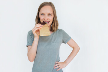 Young woman biting chocolate bar in gold foil, smiling, holding hand on waist and looking at camera. Portrait of lady isolated on white background. Front view. Tasty food and sweets concept