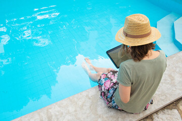 Fototapeta premium Female tourist in summer straw hat sitting at poolside, using tablet with pen, writing on screen. Outdoor shot, high angle. Vacation or wireless technology concept