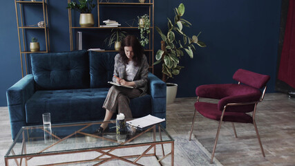 Obraz premium Young brunette woman works at home. A young woman sits on a sofa and makes notes in a notebook. Work at home during isolation. Female portrait.