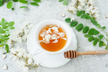 white acacia flowers tea in a white porcelain Cup on a white cement background with lush flowering branches of an acacia tree, home self-treatment, useful properties of plants, top view