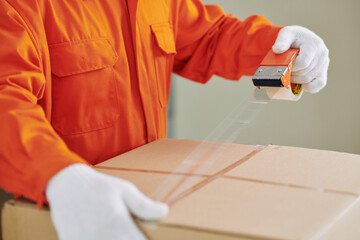 Unrecognizable house moving service worker wearing bright orange uniform closing box using tape