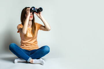 Young woman in a T-shirt and jeans sitting on the floor looks through binoculars on a light background. Banner