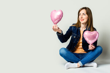 Young happy woman sitting cross-legged, dressed in jeans and jacket holding air balloons in the form of heart