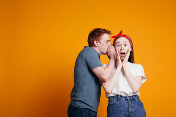 Red-haired guy whispers in ear of his girlfriend and she is very surprised at surprise. Emotional studio shot on orange background