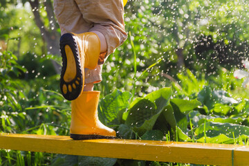 A child in yellow rubber boots stands on a bench in the rain. Feet in boots close-up. Funny kid. An...