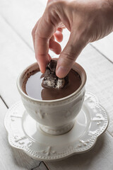 Hand dipping a chocolate Biscotti or Cantuccini into a cup of chocolate, on a white wooden table.