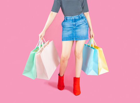 Sale and consumerism concept-Part body, beautiful female in denim short skirt, slender legs in red boots. Sexy woman holding a paper shopping bags, isolated on pink background