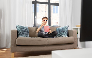 people and childhood concept - boy in 3d movie glasses eating popcorn from striped bucket and watching tv at home