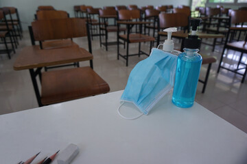 Medical face mask and alcohol hand wash gel on the teacher 's desk in front of the classroom with wood lecture chairs in the empty classroom. Concept during the Corona virus disease COVID-19 outbreak.