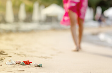 blurred woman walking on sand privat beach in pink dress, focus on shells and starfish in front, travel concept