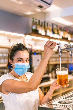 Photo session with a waitress with a face mask in a bar. New normality, security measures after the coronavirus pandemic, social distance, covid-19. Pouring a glass of pole beer and looking smiling