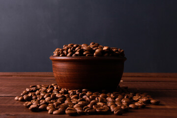 Coffee beans in clay bowl on brown wooden table against dark blue wall background. Closeup, copy space, side view. Arabica seeds, roast, caffeine concept