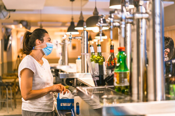 Photo session with a waitress with a face mask in a bar. New normality, security measures after the coronavirus pandemic, social distance, covid-19. Talking to a customer on the other side of the bar