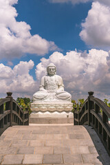 the solemn white statue of Buddha sitting on a high pedestal behind is a clear blue sky with white clouds