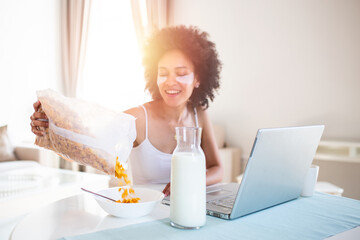 Close up smiling young mixed race woman pours corn flakes in plate with milk. The girl has a healthy breakfast on stylish cozy home at the morning while checking her email on laptop.