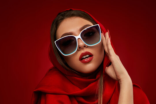 Portrait of chic brunette sexy girl with luxurious make-up in white sunglasses and red headscarf on red background