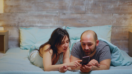 Wife in pajamas laughing while her husband is showing something on phone in the bedroom