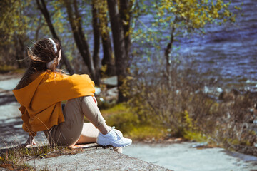A girl in a yellow sweater is sitting on the edge, listening to music with white headphones and...