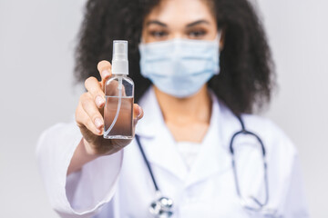 Young african american woman doctor in medical uniform and protective mask using hand sanitizer gel, isolated on grey background. Hand hygiene and antibacterial protection.