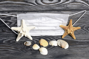 Starfish and shells are scattered on pine boards painted in black and white. Nearby is a medical mask. Holiday at sea 2020.