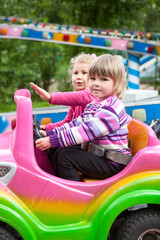 Fototapeta na wymiar Two happy little girls having fun in outdoor amusement park with riding in car on carousel, driving with steering wheel
