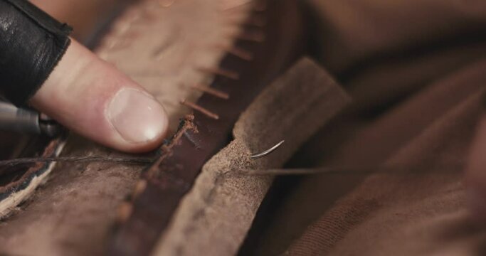 Traditional bespoke shoemaker inserts needle using awl tighten knot sewing stitching leather upper to insole on wooden shoe last with thread gloves on hands unrecognizable macro close up