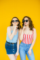 Image of two fashion women wearing girlish clothes posing at camera in stylish sunglasses isolated over yellow background