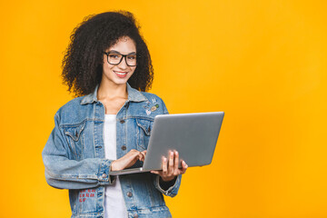 Young african american black positive cool lady with curly hair using laptop and smiling isolated over yellow background.