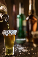 An alcoholic drink at the bar counter is poured into a tequila glass with salt at the edges, in the background are different bottles of alcohol, shallow depth of field, selective focus. The concept of