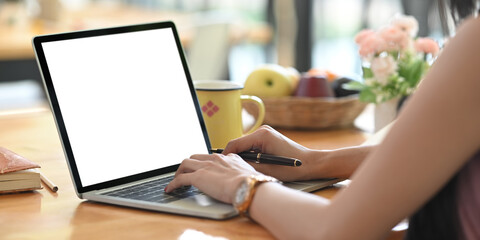 A woman is typing on a white blank screen computer laptop at the wooden working desk.