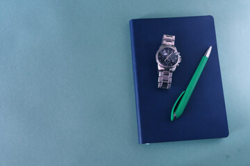 Blue diary with a wristwatch and a ballpoint pen on a green background. Doing business and management. Place for text