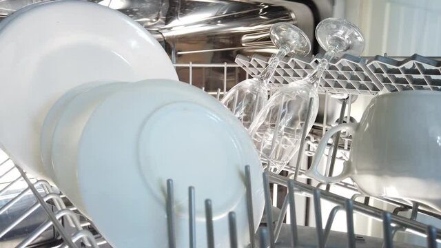 Opening dishwasher unloading dishes. Female hands pull out clean washed glass goblets and plates, the camera inside the dishwasher moves in sync with the opening