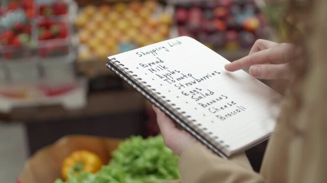 Rear view of unrecognizable woman standing in grocery marker, holding notepad with shopping list and analyzing it