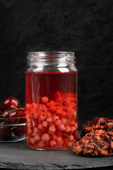 Transparent jar full of red currant compote staying on stony serving platter together with ripe cherries and oatmeal cookies
