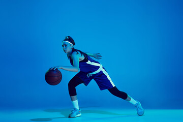 Power of youth. Young caucasian female basketball player training, prcticing with ball isolated on blue background in neon light. Concept of sport, movement, energy and dynamic, healthy lifestyle.