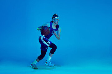 Desire to win. Young caucasian female basketball player training, prcticing with ball isolated on blue background in neon light. Concept of sport, movement, energy and dynamic, healthy lifestyle.