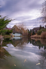 Obraz premium Old European style houses on the pond surrounded with threes and bushes. Moody sky and reflections in the pond. Vintage feel.