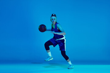 Goal achievement. Young caucasian female basketball player training, prcticing with ball isolated on blue background in neon light. Concept of sport, movement, energy and dynamic, healthy lifestyle.