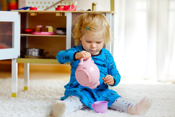 Adorable cute little toddler girl playing with toy kitchen Happy healthy baby child having fun with role game, playing with crockery teapot at home or nursery. Active daughter with toy.