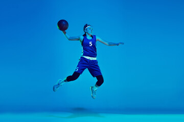 Naklejka na ściany i meble In high jump. Young caucasian female basketball player training, prcticing with ball isolated on blue background in neon light. Concept of sport, movement, energy and dynamic, healthy lifestyle.