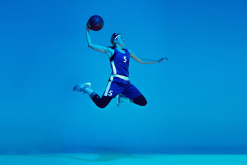 Fototapeta na wymiar In high jump. Young caucasian female basketball player training, prcticing with ball isolated on blue background in neon light. Concept of sport, movement, energy and dynamic, healthy lifestyle.