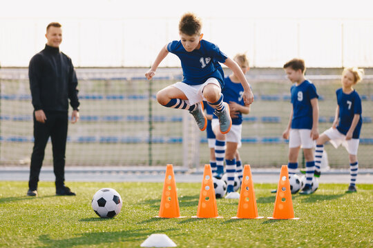 Soccer School Training Unit. Football Boys In Team On Practice Session With Youth Coach. Player Jumping Over Training Cones On The Grass Field. Speed And Agility Soccer Training