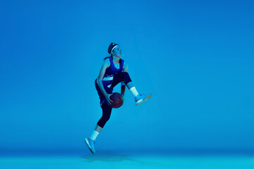 Fototapeta na wymiar In high flight. Young caucasian female basketball player training, prcticing with ball isolated on blue background in neon light. Concept of sport, movement, energy and dynamic, healthy lifestyle.