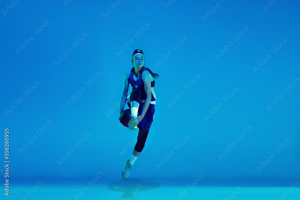 Wall mural In high flight. Young caucasian female basketball player training, prcticing with ball isolated on blue background in neon light. Concept of sport, movement, energy and dynamic, healthy lifestyle.