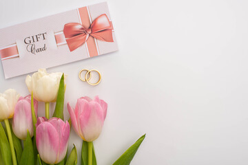Top view of gift card near flowers and wedding rings on white background