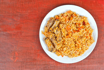 Pilaf with meat and carrots in a white plate. Rice on red plywood. Place for text and copy space near food.