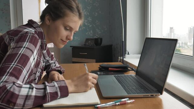 Tired european female student study with online tutor teacher, look at laptop computer and make notes, online conference, online study