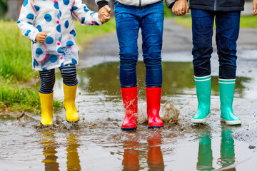 Three children, toddler girl and two kids boys wearing red, yellow and green rain boots and walking during sleet. Happy siblings jumping into puddle. Having fun outdoors, active family